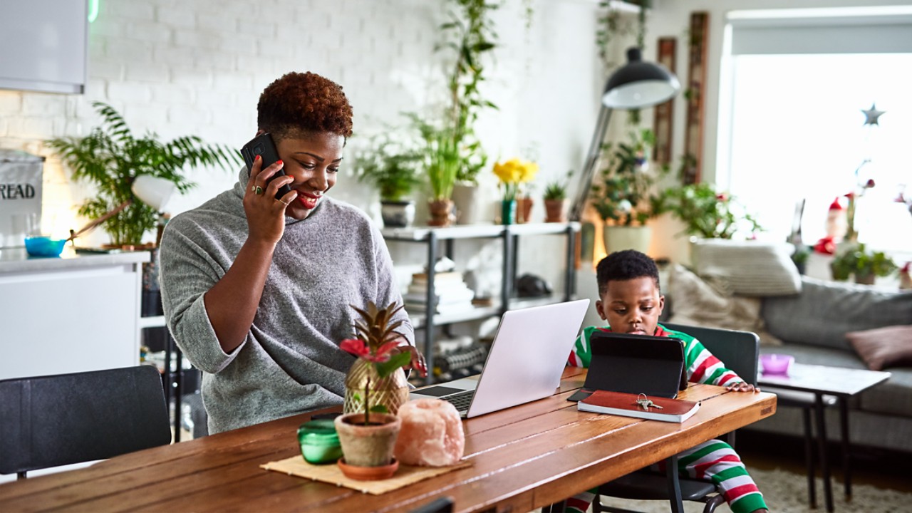 Women talks on the phone in the kitchen of her home. A laptop is open in front of her, a boy is watching a tablet beside her.